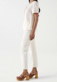 Femme en haut à manches courtes blanc cassé et pantalon, portant des sabots en bois à talons marron avec boucle et clous, se tenant de profil.