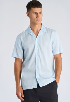 Mężczyzna ubrany w jasnoniebieską koszulę na krótki rękaw z guzikami i czarne spodnie, stoi z jedną ręką w kieszeni, patrząc na bok na tle jednolitego tła.