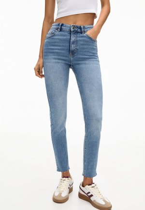 Femme portant un jean skinny taille haute bleu clair avec des ourlets bruts, des baskets blanches et un top court blanc, posant avec une main dans la poche.