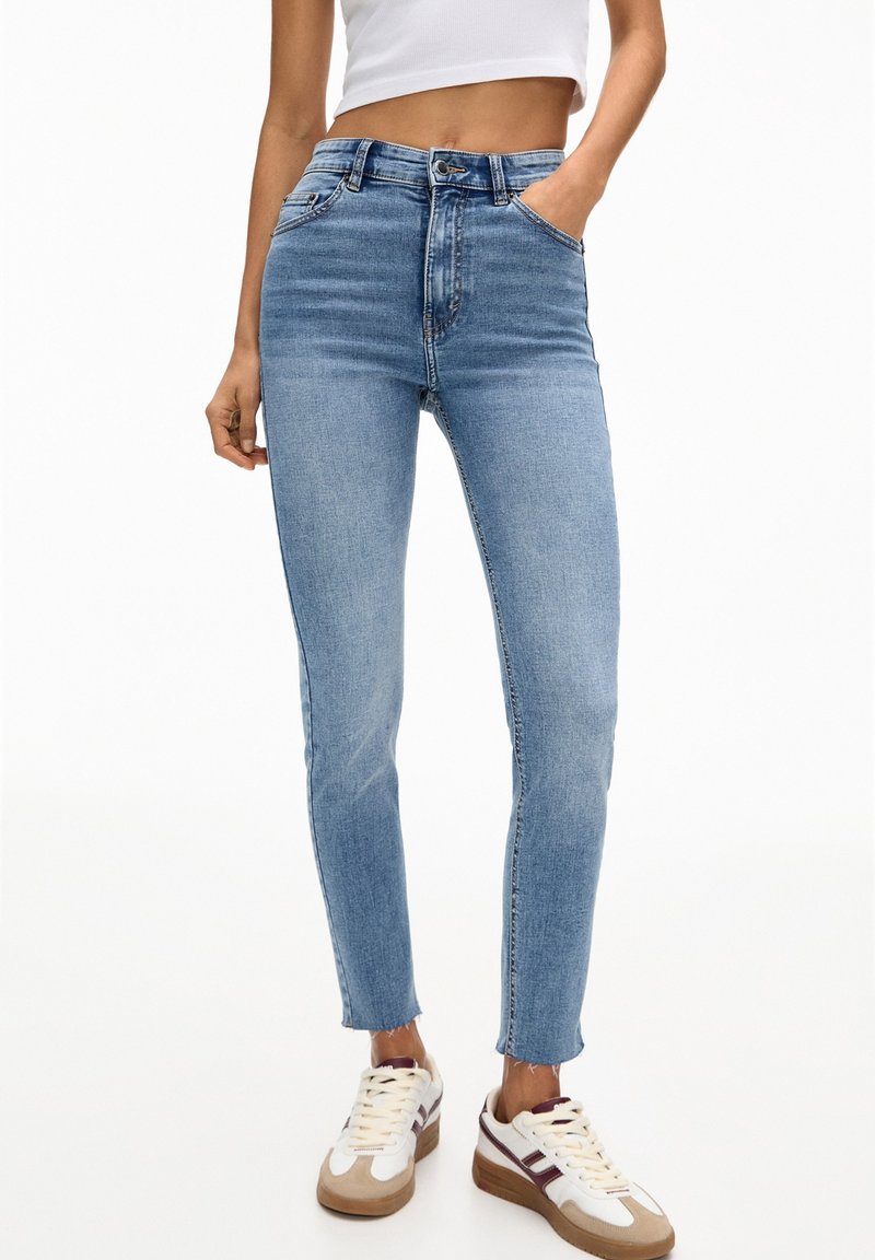 Femme portant un jean skinny taille haute bleu clair avec des ourlets bruts, des baskets blanches et un top court blanc, posant avec une main dans la poche.