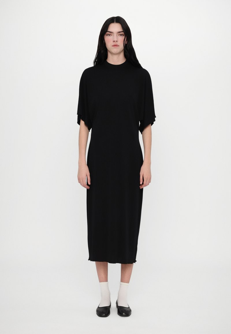 Robe noire mi-longue avec des manches courtes, en tissu lisse. Présente une coupe droite et des détails d'ourlet brut, portée avec des chaussures noires et des chaussettes blanches.
