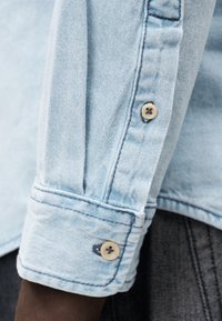 Camisa de ganga azul-claro com punho abotoado, textura macia e botões contrastantes com um detalhe de costura visível ao longo da manga.