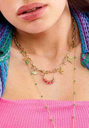 Goldkette mit verschiedenen Anhängern: einem Halbmond, einer Schildkröte, einer Muschel und grünen Perlen. Getragen über einem pinken strukturierten Oberteil.