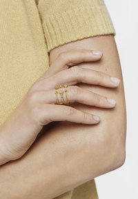 Gold rings feature varying shapes and textures, worn on a hand resting on a bare arm, against a light yellow knit background.