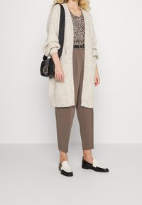 Vrouw in een beige lange gebreide cardigan, met een luipaardpatroon top, taupe broek, zwart-witte loafers en een zwarte schoudertas.