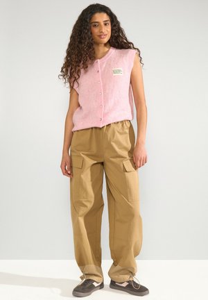 Vrouw met lang krullend haar draagt een roze mouwloos knoopgilet en losse beige cargobroeken met zwart-witte sneakers.