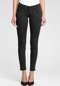 Vaqueros pitillo negros en una tela elástica con una textura suave, con bolsillos delanteros y cintura de talle medio, combinados con tacones altos negros.