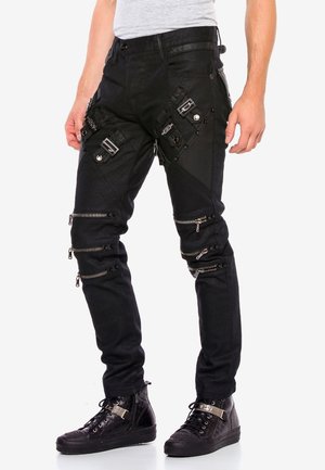 Homme portant un pantalon cargo noir avec plusieurs poches zippées, des sangles et des boucles, associé à des baskets montantes noires.