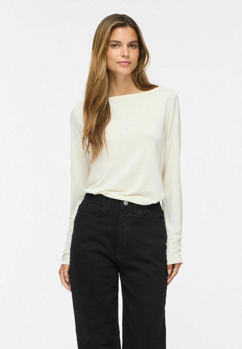 Femme aux longs cheveux bruns portant un haut blanc à manches longues et un jean noir taille haute, debout devant un fond clair uni.