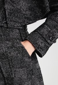 Černo-bílý kabát s potiskem houndstooth, texturovaná tkanina, velký límec, knoflíkové manžety a pásek svázaný v pase.