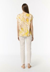 Kurzärmlige Bluse mit einem gelb- und braunfarbenen Blattmuster, kombiniert mit weißen Slim-Fit-Hosen und hellbraunen Sandalen. Glatte Textur.