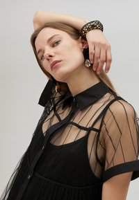 Robe noire transparente avec un col, des motifs à rayures verticales et un tissu doux au toucher. Le mannequin accessoirise avec une grande boucle d'oreille et un bracelet.