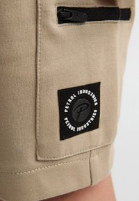 Beige bomuldsshorts med en struktureret overflade, sidelomme med lynlås og et sort mærke med teksten "Petrol Industries" i hvide accenter.