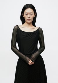 Sort bluse med lange ærmer og rund udskæring, med gennemsigtige ærmer og en blød tekstur, kombineret med et sort tekstureret skørt.