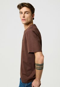 Ruskea lyhythihaainen t-paita pehmeästä kankaasta, joka näyttää yksinkertaiselta ja jossa on pieni musta logo rinnassa. Yhdistettyinä sinisiin farkkuihin.