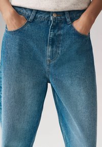 Helle blaue Jeans mit hoher Taille, großen frontalen Taschen und subtilen Nähdetails. Die Textur wirkt glatt mit leichten Verblassungen.