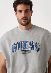 Sivi bezrukavi dukserica s velikim "GUESS" logom u plavoj i crnoj boji, uz tekst "ATHL DEPT" ispod. Pamučni materijal s mekom teksturom.