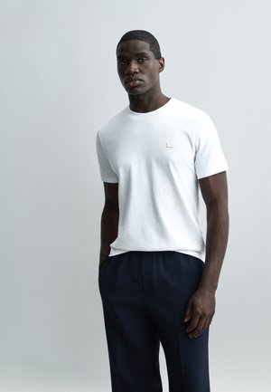 Homme noir debout, les mains dans les poches, portant un t-shirt blanc uni à manches courtes et un pantalon sombre, sur un fond gris clair.