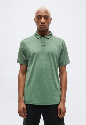 Uomo con capelli biondi corti che indossa una polo a maniche corte verde e pantaloni neri, in piedi davanti a uno sfondo chiaro e semplice.