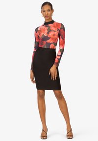 Langärmliger, taillierter Bodysuit mit floralem Muster in Rot und Orange, kombiniert mit einem schwarzen, knielangen Bleistiftrock. Das Model trägt schwarze, geschnürte High Heels.