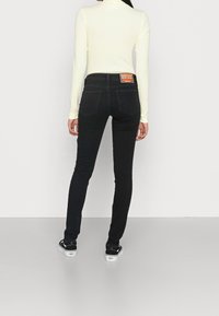 Vaqueros skinnies negros de cintura alta, con bolsillos traseros y una etiqueta de parche de cuero en la cinturilla, combinados con una blusa de manga larga de color amarillo claro.