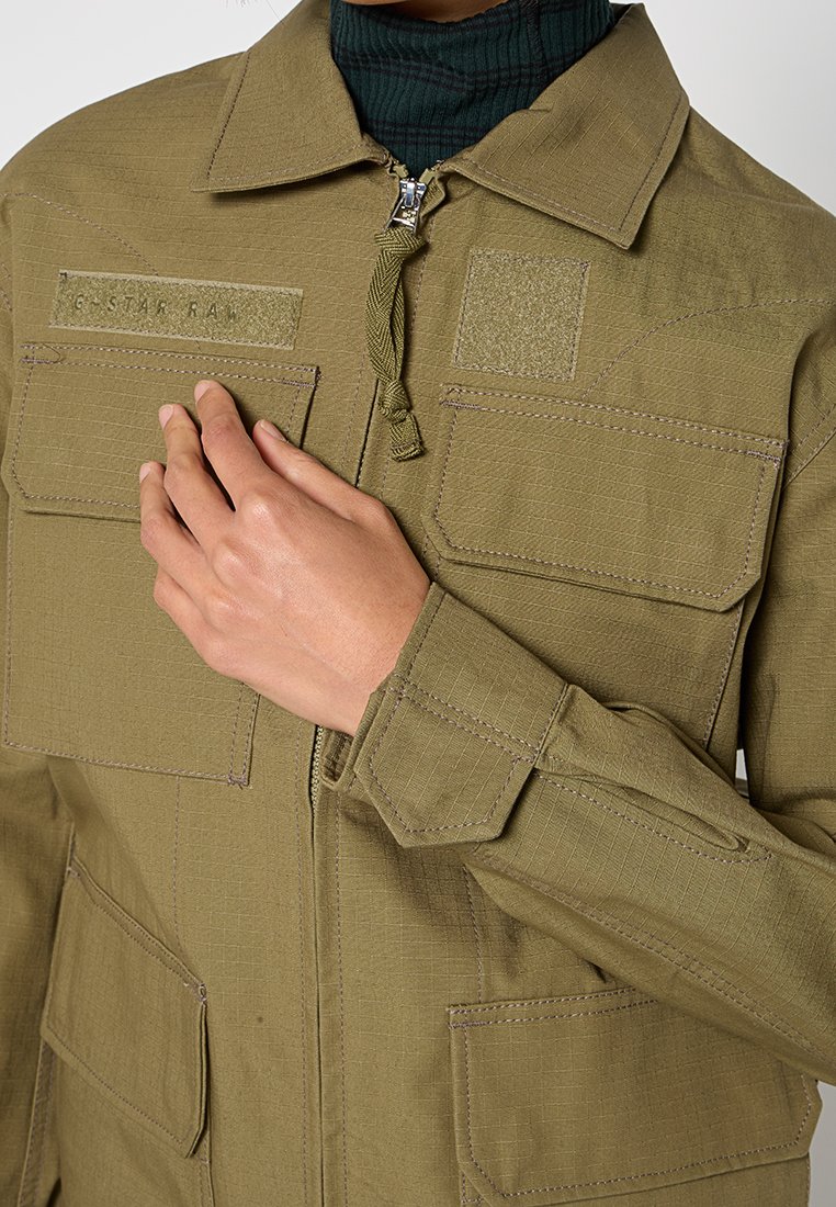 Olivgrüne Jacke mit Reißverschluss, vier Fronttaschen, Patches und strukturiertem Stoff. Sie hat einen hohen Kragen und umgeschlagene Ärmel.