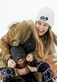 White knit beanie with black text and owl graphic, worn by a woman with long hair, hugging a man in a dark outfit, snow falling around them.