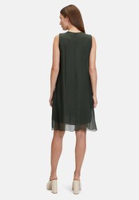 Ärmelloses, dunkelgrünes Kleid mit lockerer Passform, seidenähnlicher Textur und einer Mittelnaht hinten. Kombiniert mit hellen Plateau-Heels.