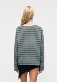 Γυναίκα με μακριά ξανθά μαλλιά, που φοράει ένα μπλε και καφέ ριγέ πουλόβερ και μια ασύμμετρη μαύρη φούστα, στέκεται μπροστά σε ένα απλό λευκό φόντο.