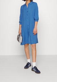 Abito blu con bottoni, maniche a tre quarti, colletto e vestibilità comoda. Abbinato a scarpe sportive scure e una borsa a tracolla argentata.