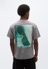 Γκρι βαμβακερό T-shirt με μεγάλο γραφικό τύπωμα γυαλιών ηλίου στην πλάτη, με ξεθωριασμένο πράσινο φόντο και υφή υφάσματος.