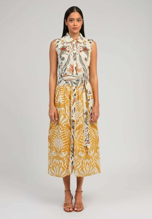 Jeune femme debout, portant une robe midi sans manches à col, avec un motif floral blanc et jaune et des feuilles, ainsi que des sandales à bride cheville.