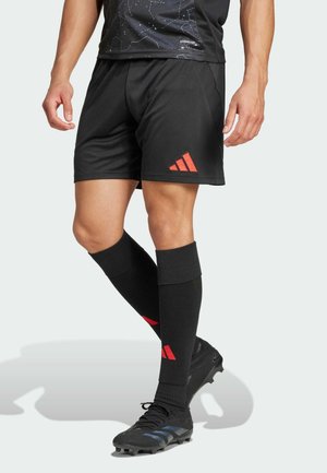 Mann trägt schwarze Sportshorts, schwarze knielange Socken mit roten Adidas-Logos und schwarze Fußballschuhe vor einfachem Hintergrund.