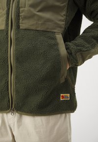 Jachetă din polar verde măslin cu un panou din material neted, închidere cu fermoar, buzunare laterale și o aplicație cu logo. Suprafață texturată și croi lejer.
