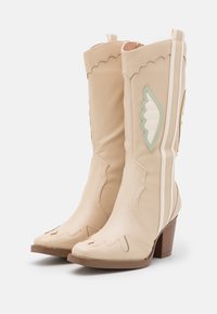 Botas de vaquero de cuero beige con bordes superiores ondulados, costuras decorativas, acentos turquesa y un tacón de madera apilada.