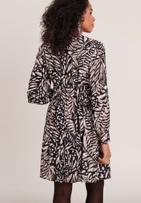 Robe à manches longues avec un motif de feuilles noir et beige, dotée d'une taille élastique et de détails froncés ; le tissu semble léger et texturé.
