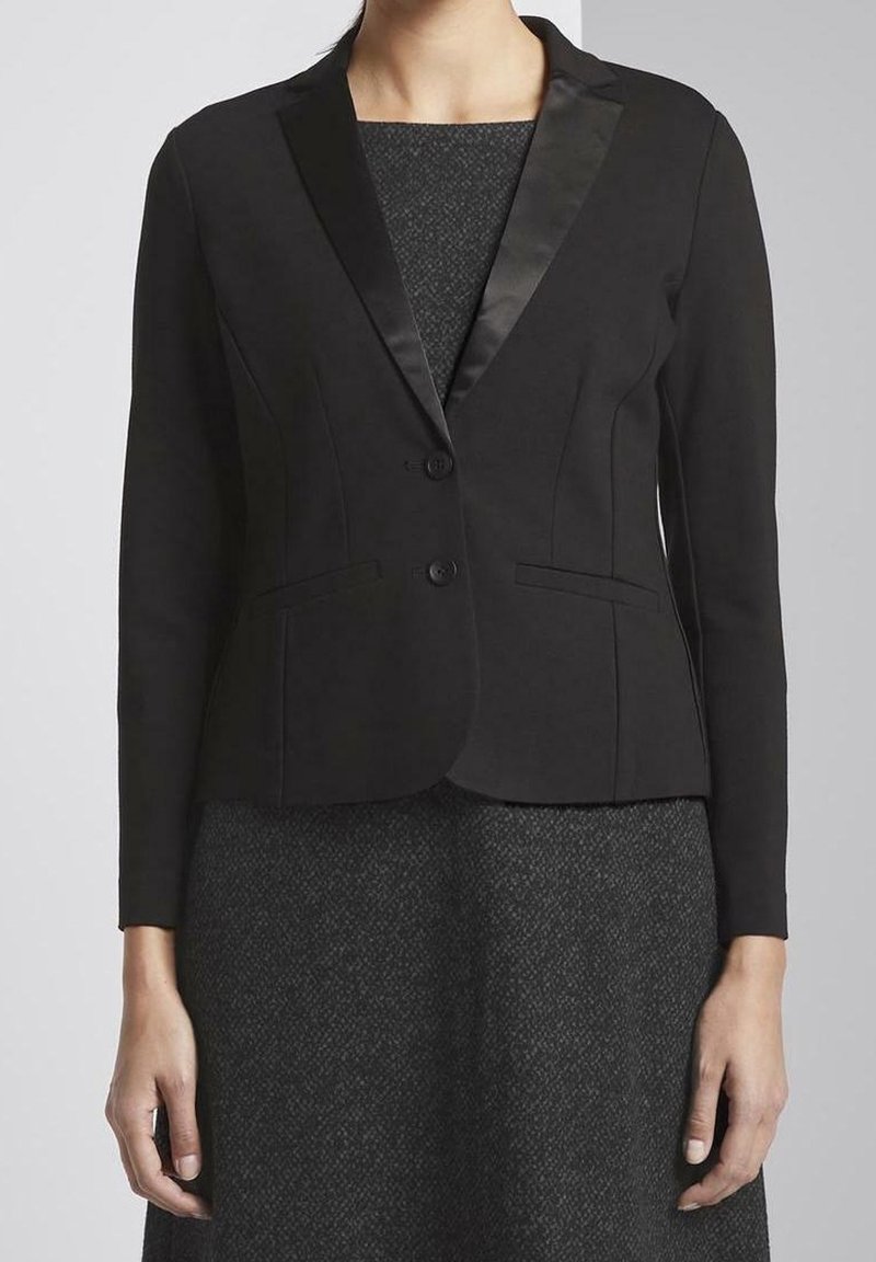 Blazer nero con rever in satin, chiusura a un bottone e due tasche frontali. Vestibilità su misura e maniche lunghe. Indossato sopra un vestito grigio texturizzato.