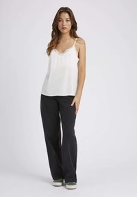 Camisole en soie blanche avec des bordures en dentelle, associée à un pantalon noir évasé et des baskets en daim vert. Le modèle se tient avec une expression neutre.