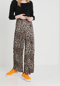 Leopardikuosiset leveälahkeiset housut teksturoidusta kankaasta, joissa on korkea vyötärö ja sivuaukot, yhdistettynä kirkkaan oransseihin tennareihin.