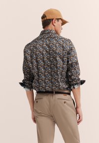 Braunblumiges Hemd mit blauen und weißen Akzenten, hochgekrempelte Ärmel, kombiniert mit hellbeigen Hosen und einer braunen Kappe. Lässiger Schnitt.