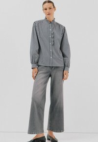 Vrouw in een zwart-witte geblokte blouse met knopen en ruffles, met brede grijze jeans, staande tegen een effen witte achtergrond.