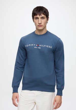 Νέος άνδρας φοράει μπλε φούτερ Tommy Hilfiger με λογότυπο και άσπρο παντελόνι, στέκεται σε απλό φωτεινό φόντο.