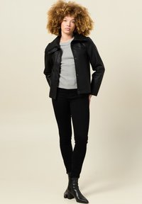 Giacca di pelle nera con colletto classico, chiusura a bottoni e maniche lunghe, indossata sopra un maglione grigio e abbinata a jeans neri e stivaletti.