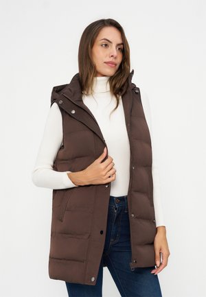 Femme portant une veste matelassée sans manches marron à capuche par-dessus un pull à col roulé blanc et un jean bleu, debout devant un fond uni.