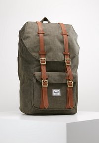 Mochila de tela verde con correas de cuero marrón y hebillas doradas, bolsillo frontal y una etiqueta blanca de la marca Herschel.