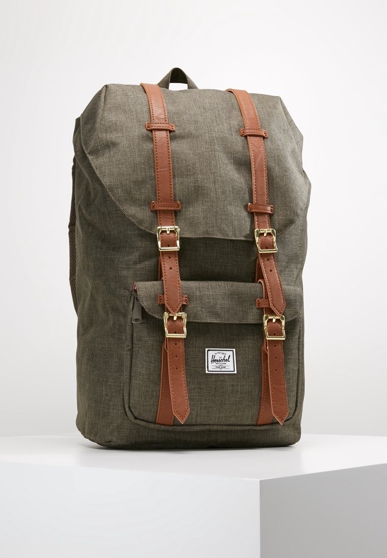 Mochila de tela verde con correas de cuero marrón y hebillas doradas, bolsillo frontal y una etiqueta blanca de la marca Herschel.