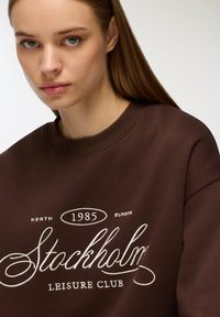Felpa marrone con la scritta "Stockholm Leisure Club" ricamata in bianco, con scollatura a costine e vestibilità ampia.