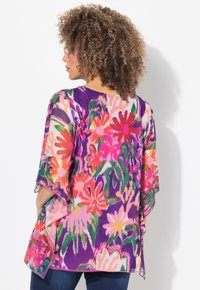 Blusa viola trasparente con motivi floreali in rosa, arancione e verde. Presenta maniche larghe e una vestibilità ampia, l'orlo cade appena sotto i fianchi.
