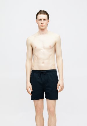 Fiatal férfi áll, előre néz, ing nélkül, fekete zsinóros fürdőnadrágot visel, egyszínű világos háttér előtt.