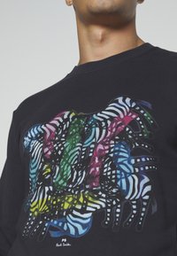 Maglietta nera con motivo astratto di zebra colorato in blu, verde, giallo e rosa sul petto, indossata da una persona vista dal collo in giù.